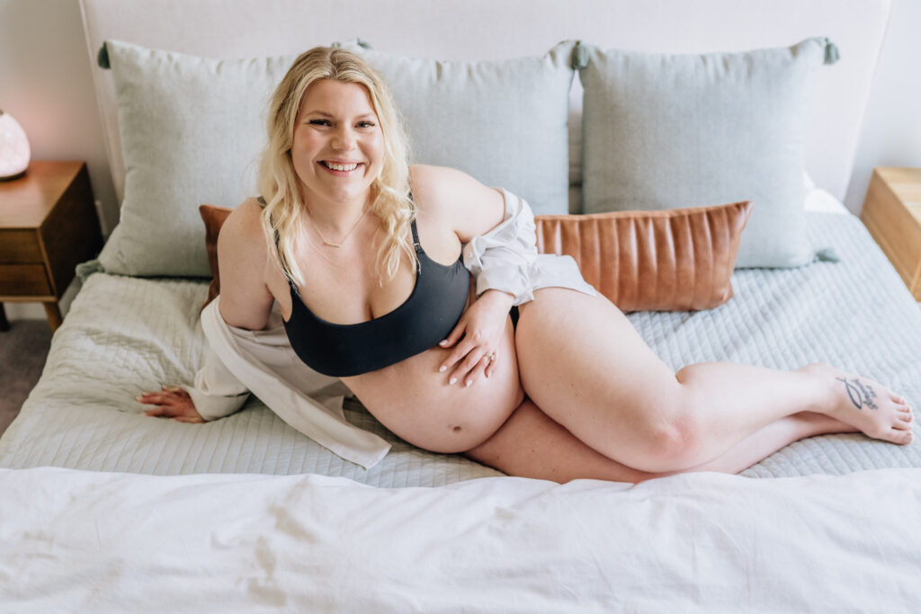 A pregnant woman, smiling and looking at the camera, is reclining on a bed with light green pillows and a brown cushion. She is wearing a black bra and has a white shirt draped over her shoulder, with a tattoo visible on her right foot. The ambiance is calm and cozy, with a bedside table and a lamp in the background.