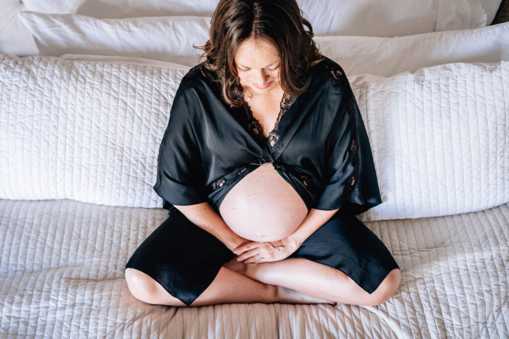 A pregnant woman is seated cross-legged on a bed, looking down at her bare belly. She is wearing a black robe with lace details, which is open at the front to reveal her pregnant stomach. The bed is covered with white, quilted bedding, creating a soft and serene atmosphere that highlights the woman's contemplative and peaceful expression.