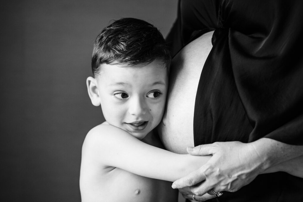 A black and white image of a young boy hugging a pregnant woman's belly. The boy looks to the side with a curious and excited expression, while the woman, draped in a dark garment, gently holds him close. The intimate moment captures the bond between the child and the expected sibling.