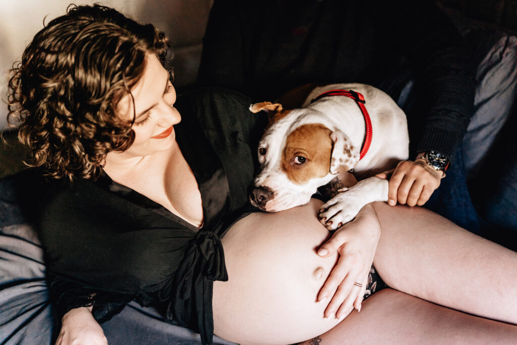 A pregnant woman reclines on a bed, with a dog resting its head gently on her bare belly. She is smiling down at the dog, wearing a black robe tied at the front. The dog, wearing a red collar, looks up with a calm and affectionate expression. The scene is warm and tender, highlighting the bond between the woman and her pet.