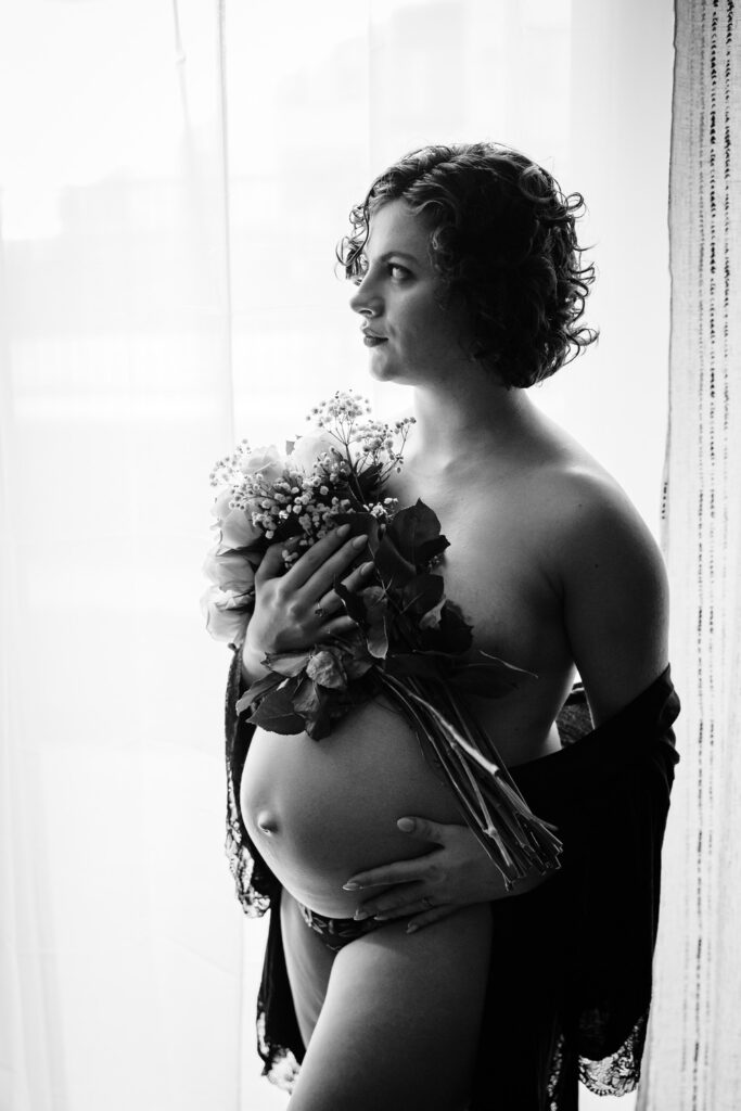 A black and white image of a pregnant woman standing in front of a sheer curtain, holding a bouquet of flowers close to her chest. She is draped in a dark lace garment, with her gaze directed thoughtfully to the side. The soft lighting creates a serene and intimate atmosphere, highlighting her pregnancy and the floral arrangement.