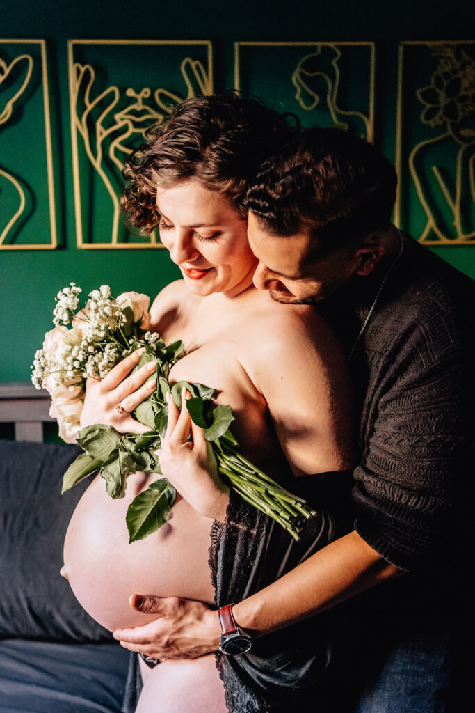 A pregnant woman, holding a bouquet of roses and baby's breath, is embraced from behind by her partner. She is smiling softly, and her partner is gently kissing her shoulder. They are standing in front of a green wall adorned with minimalist line art. The lighting highlights the intimacy and warmth of the moment.