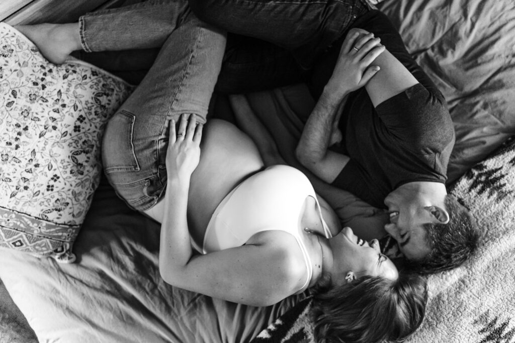 A black and white image of a pregnant woman and her partner lying on a bed, facing each other and smiling. The woman is wearing a white bra and jeans, while the man is in a dark t-shirt and jeans. They are cuddled close, with the woman's hand resting on her partner's chest and his arm wrapped around her. The bed is adorned with a patterned pillow and a textured blanket, adding to the cozy atmosphere.