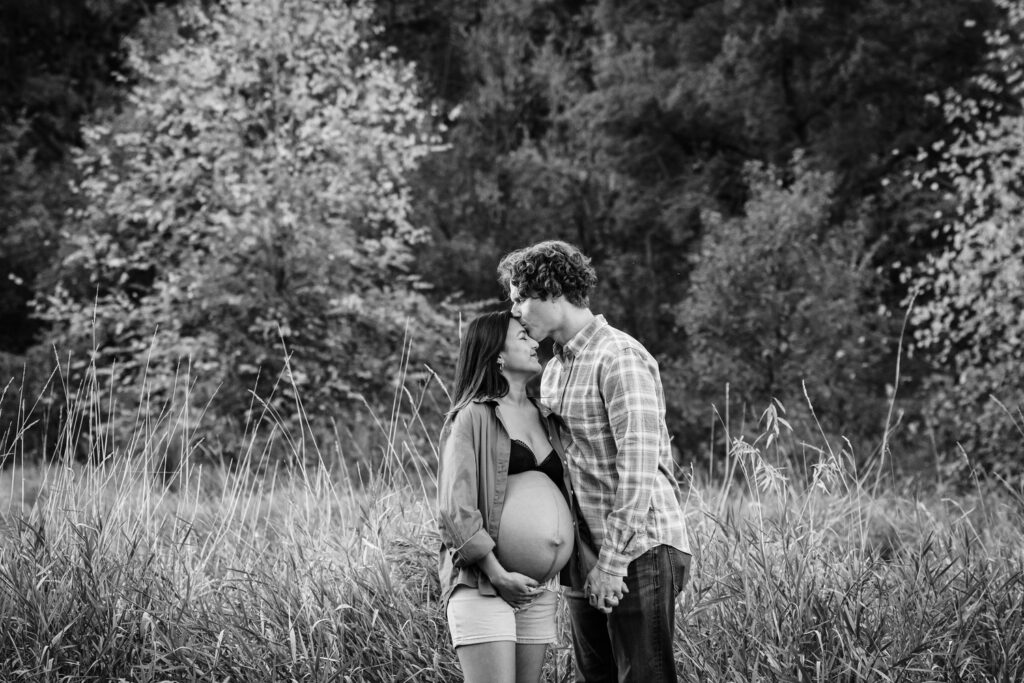 A black and white image of a pregnant woman and her partner standing in a field with tall grass. The woman is wearing a button-up shirt over a bra and shorts, and her partner is in a plaid shirt and jeans. They are holding hands, with the man gently kissing her forehead, creating a tender and loving moment. Trees form the backdrop, adding a natural and serene ambiance.