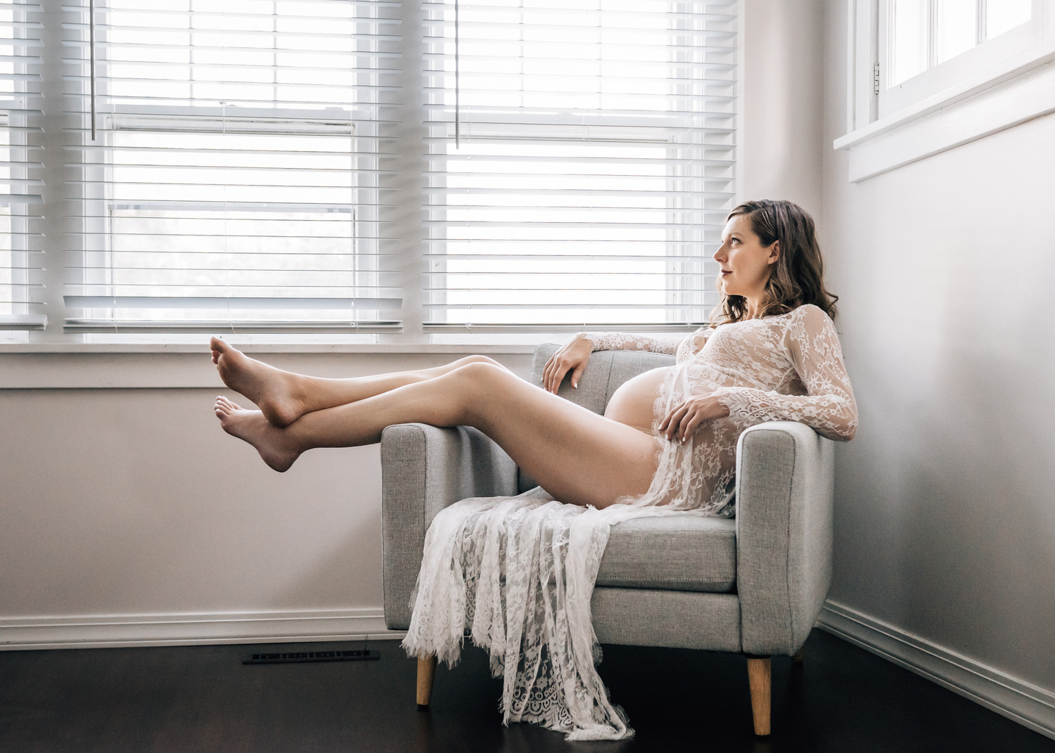 A pregnant woman is lounging in a light gray armchair, with her legs stretched out and resting on the armrest. She is wearing a delicate, lace robe that drapes elegantly over the chair. The room is bright with natural light streaming in through the white blinds, creating a serene and relaxed atmosphere.