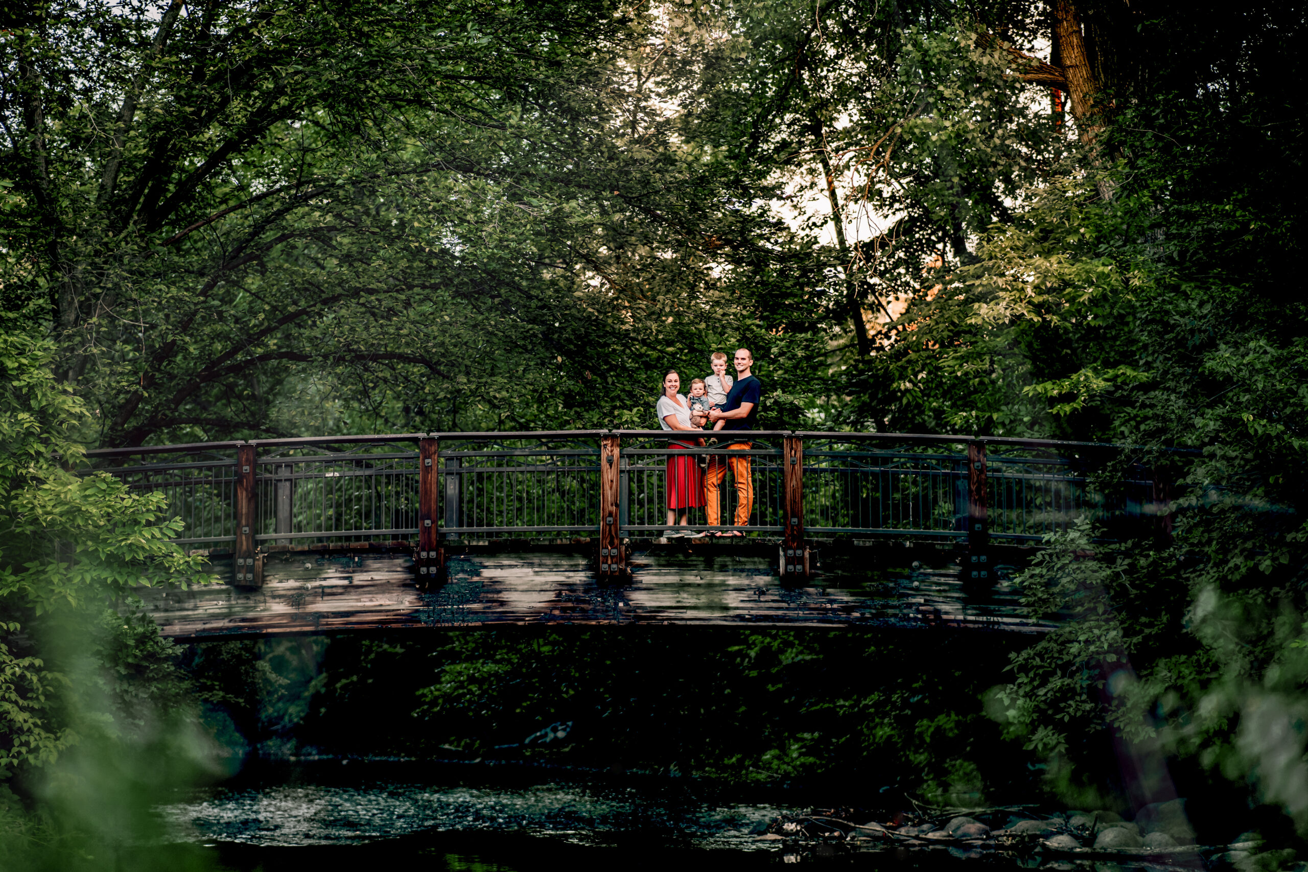 family standing on a bridge together surrounded by trees with a river 