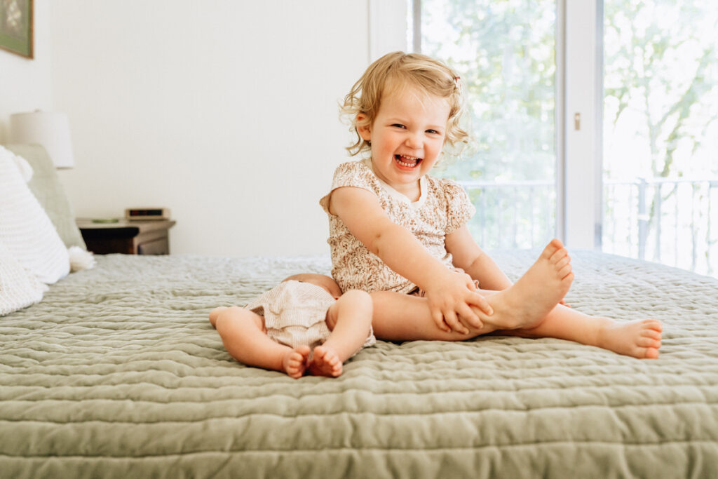 a toddler sitting on a bed smiling with a newborn baby laying next to them 
