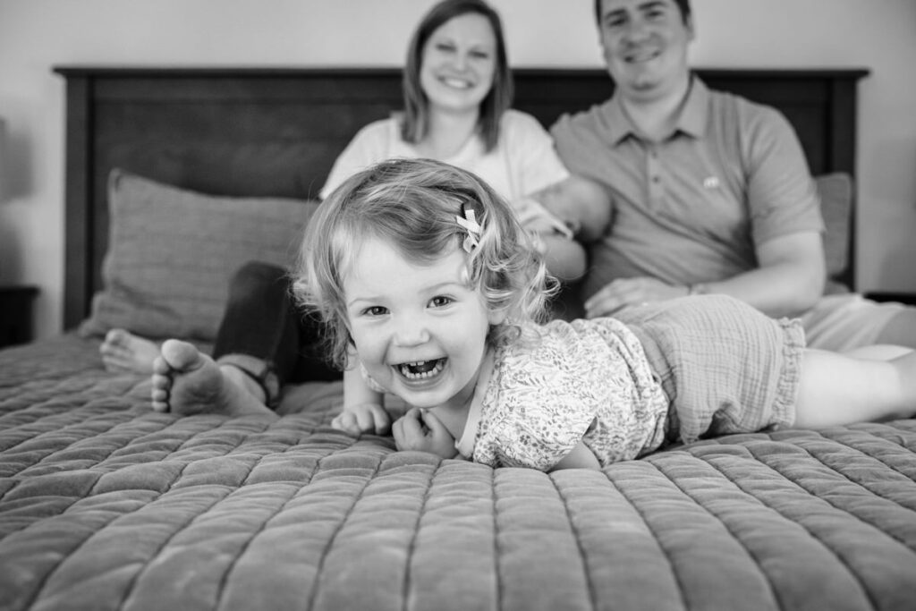 a young child lying on a bed, smiling directly at the camera while parents sit in the background holding a newborn