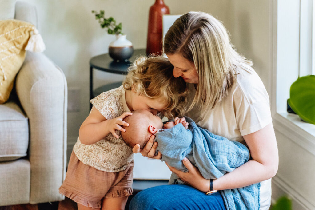 a mother holding a newborn baby while a young child kisses the baby on the head