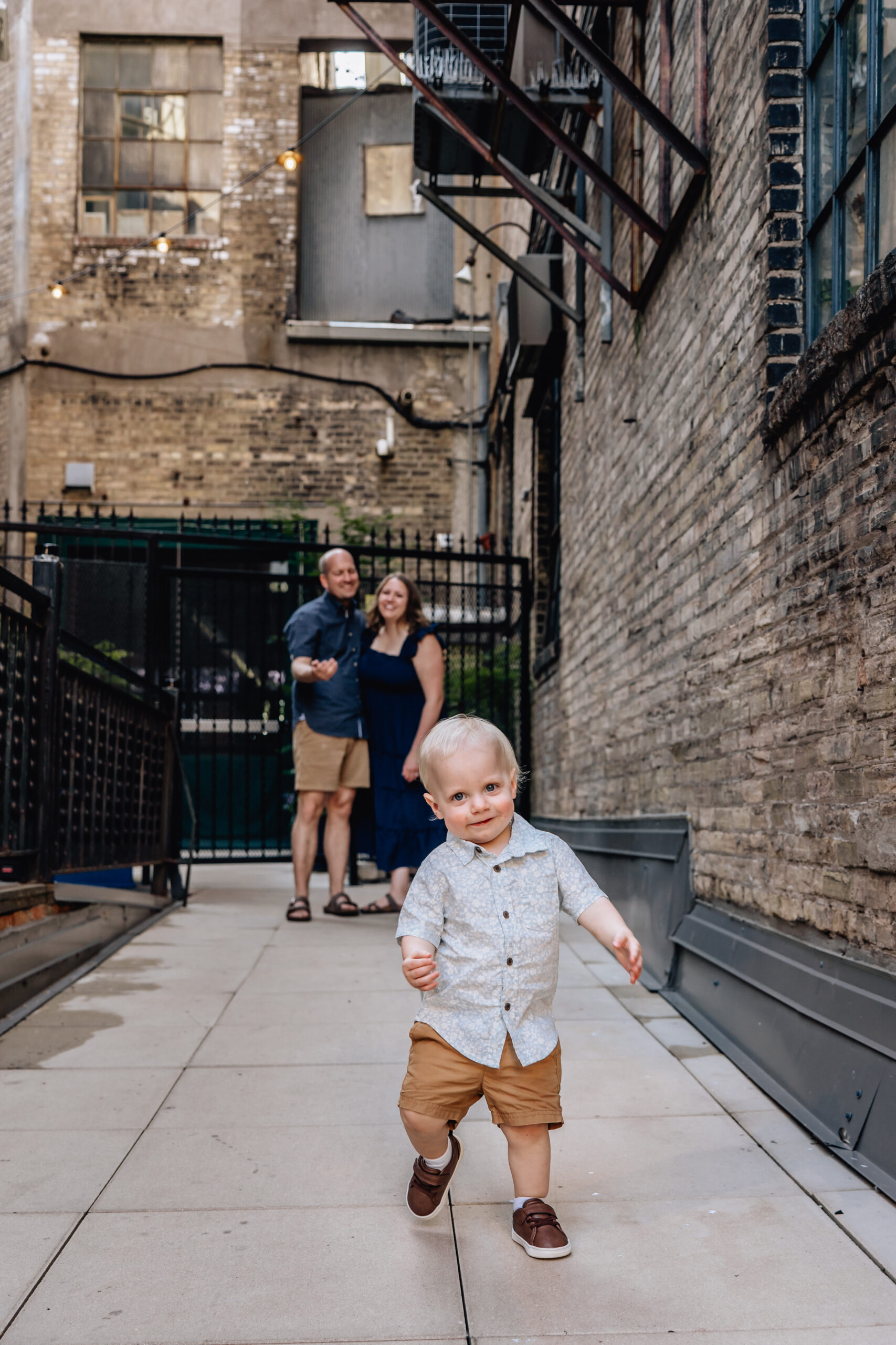 toddler walking toward the camera while mom and dad watch smiling in the background 