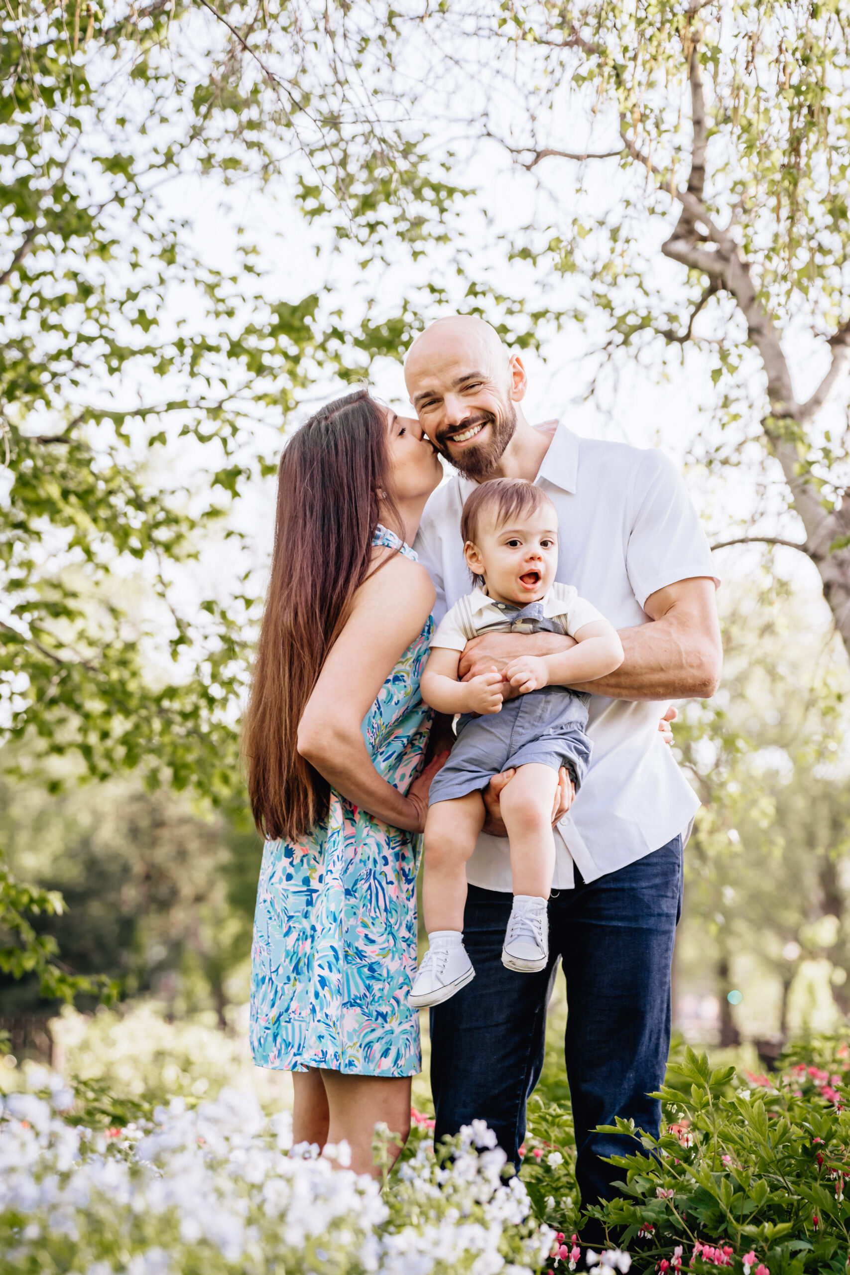 Mother kissing her husband on the cheek while her husband holds their toddler son in his hands