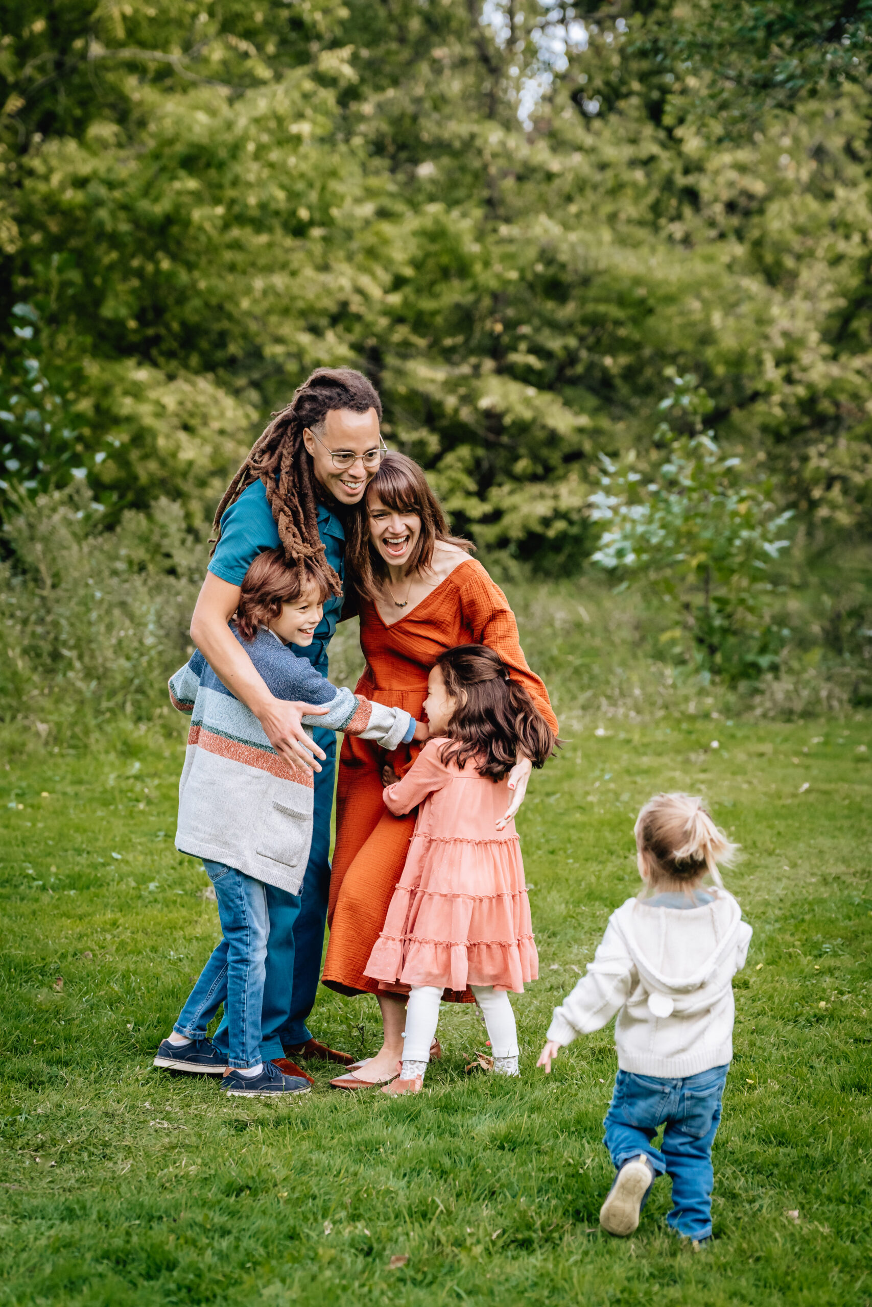 father and mother hugging their daughter and son while other toddler daughter runs through the grass