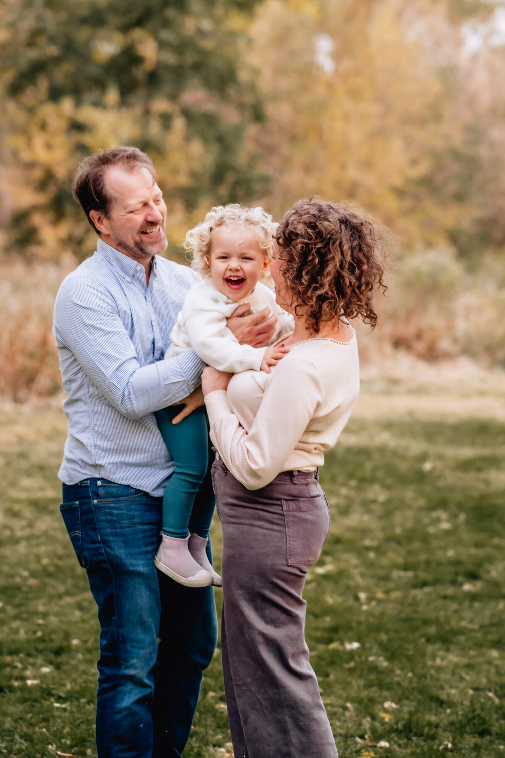 mother and father laughing and looking at their toddle daughter who is giggling 