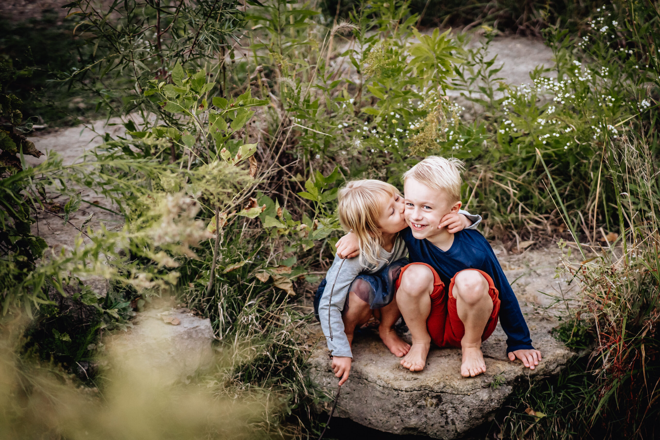 young brother and sister sitting on the rocks - sister is giving her brother a kiss on the cheek 