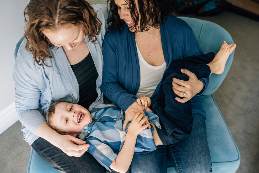 two mothers sitting on a chair with their child lying across their laps, all smiling