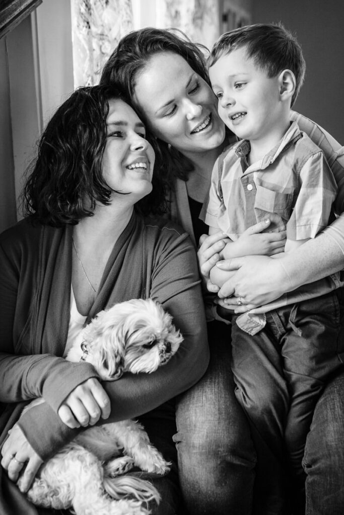 two mothers sitting with their child and a small dog by a window, smiling