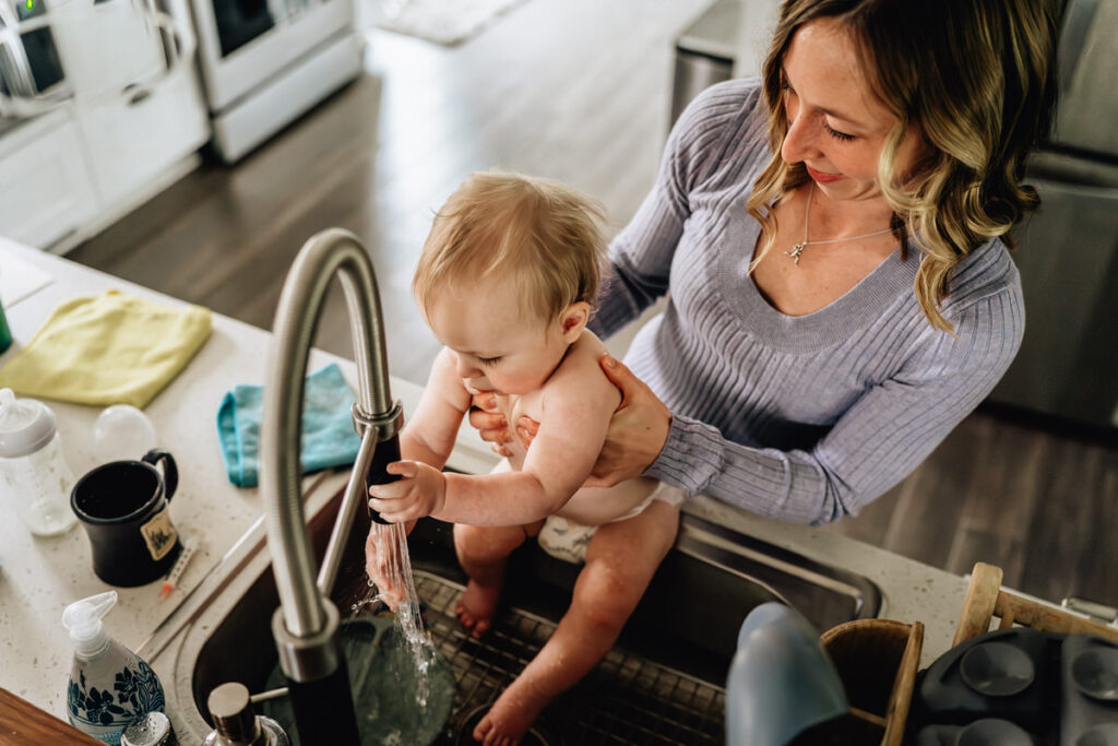 a mother holding a baby over a kitchen sink, letting the baby play with running water from the faucet