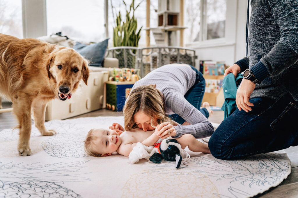 a mother kissing a baby on the belly while the baby lies on a play mat with a dog nearby