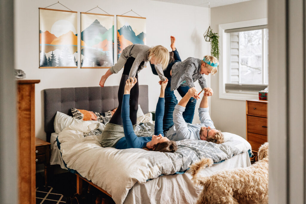 a family playing on a bed, with parents holding their children in the air, and a dog standing nearby