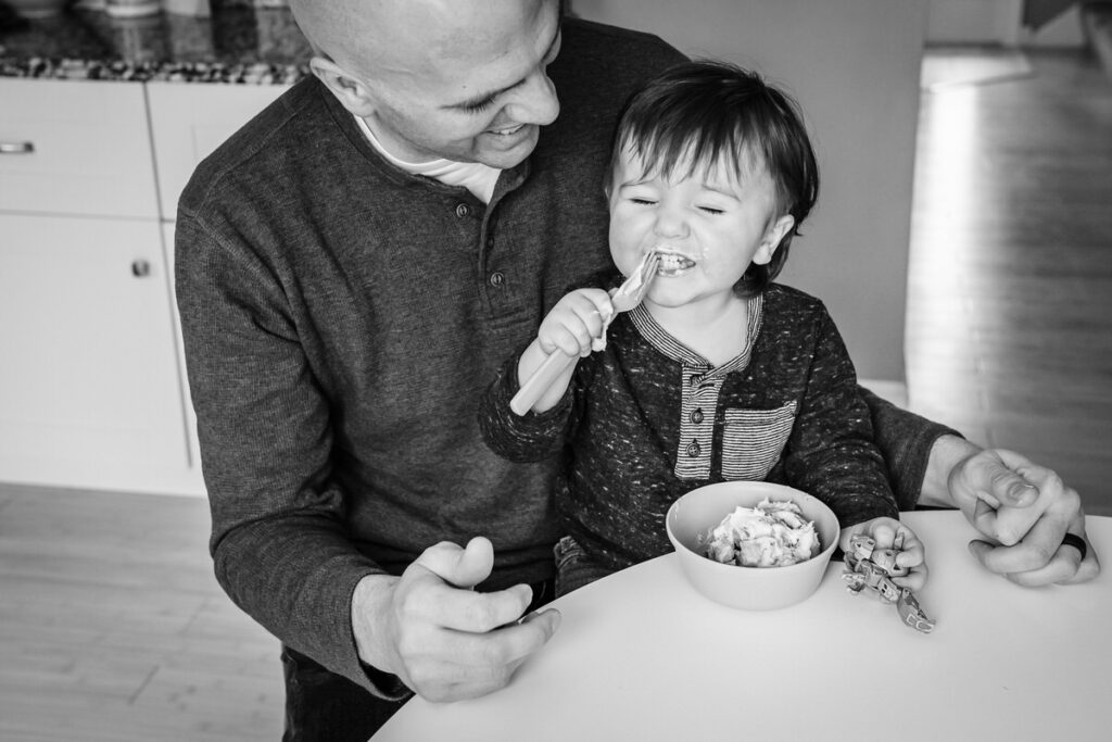 a father sitting with his child at a kitchen table, helping them eat from a bowl with a spoon