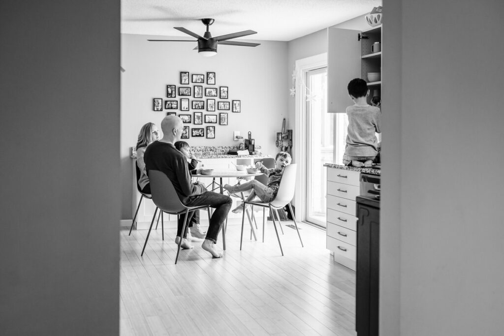 a family sitting at a kitchen table, having a meal together while children play nearby