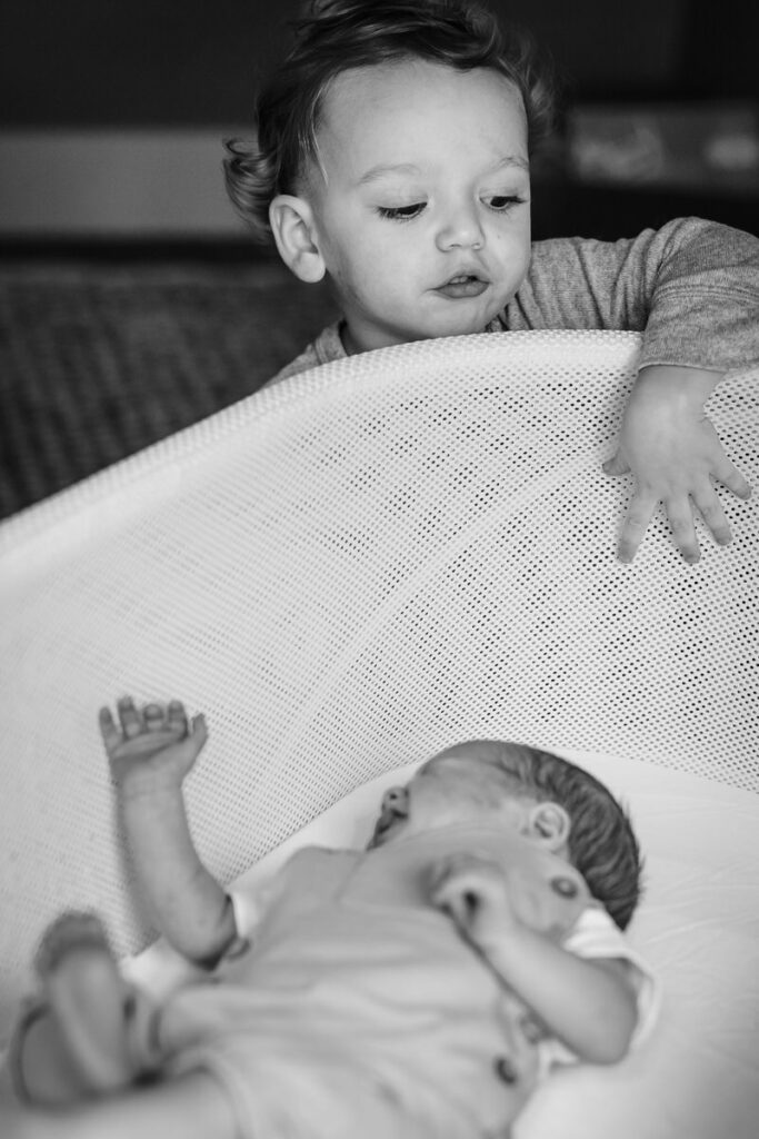 a young toddler looking over the edge of a bassinet at a newborn baby