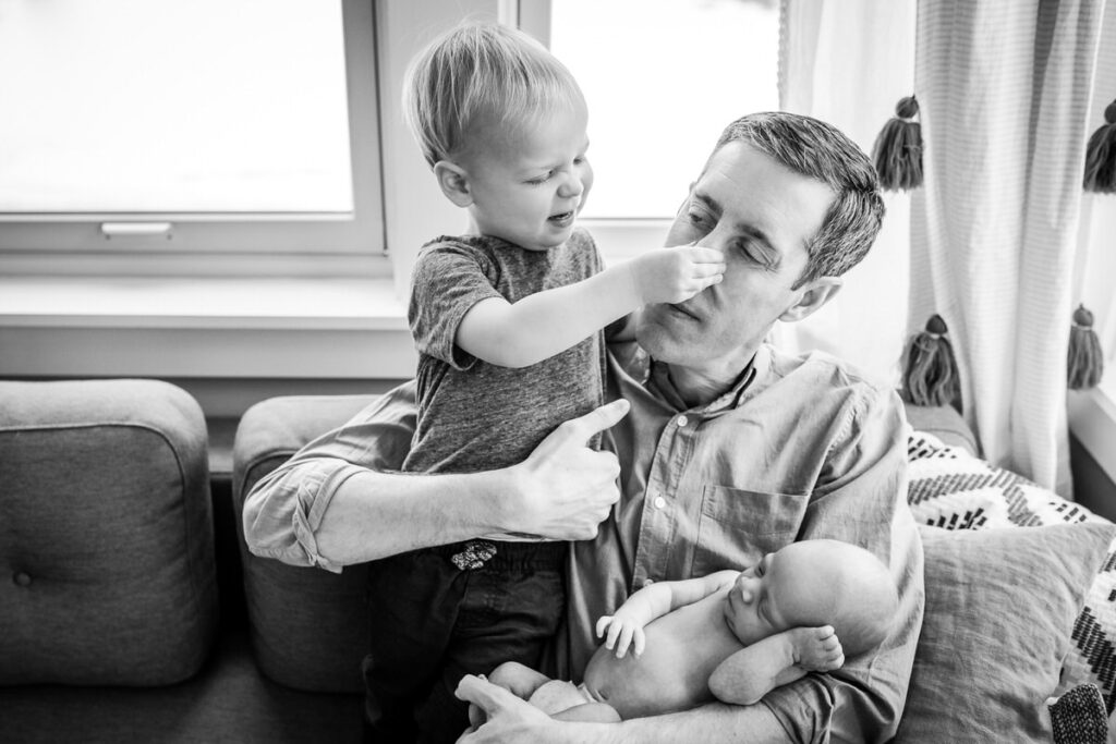 a father holding a newborn baby while his older child grabs his nose