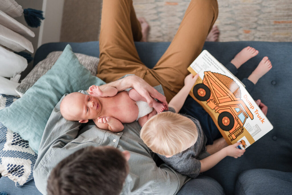 a father holding a newborn baby and reading a book about dump trucks with his older child