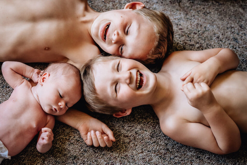 two young boys lying on the floor, laughing and smiling while the newborn sleeps beside them