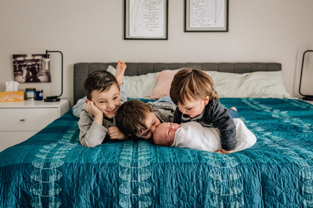 three young boys lying on a bed, two of them kissing and cuddling their newborn sibling