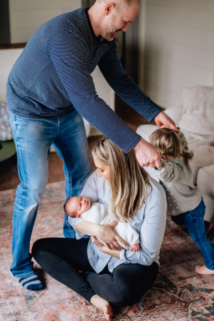 a mother sitting on the floor cradling a newborn baby while the father plays with an older child 