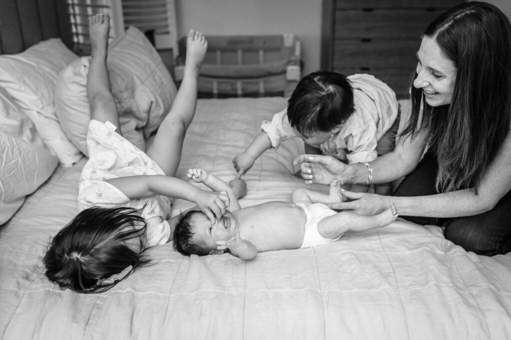 two young children playing on a bed with their newborn sibling, while their mother watches and smiles