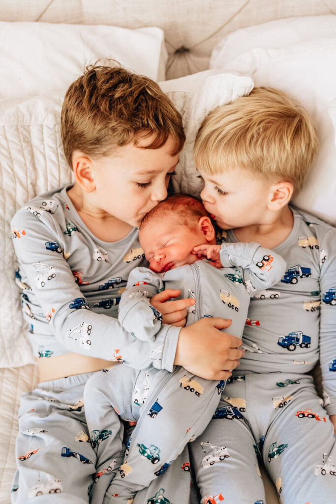 Two young boys in matching pajamas holding a newborn sibling; one boy kisses the baby on the head while the other watches