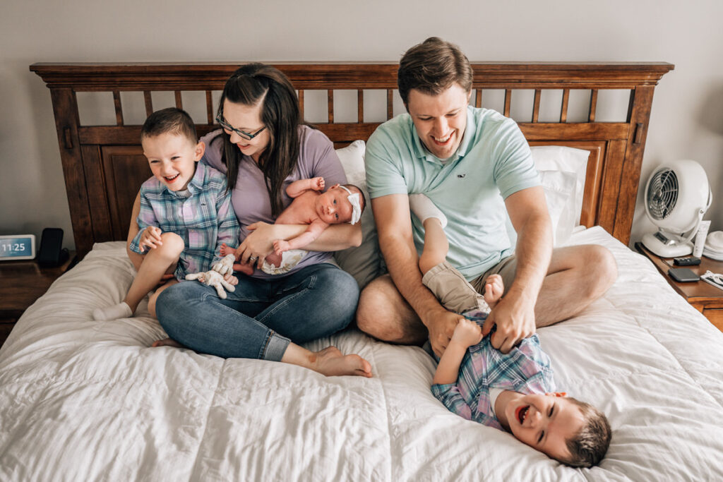 a family sitting on a bed, with parents holding a newborn baby and playfully interacting with their older children