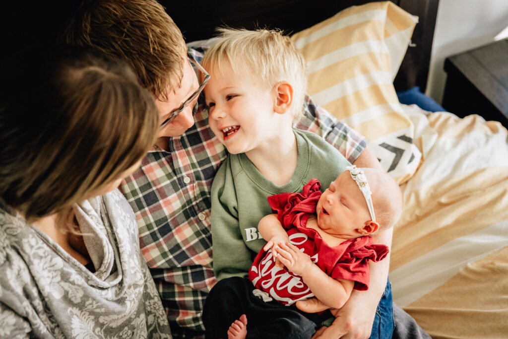 a family gathered on a bed, with parents looking lovingly at their newborn baby while an older child smiles brightly