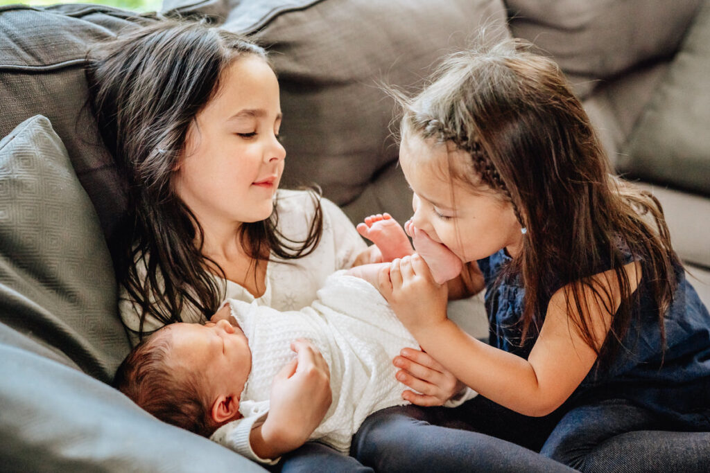 two young girls sitting on a couch with one holding a newborn baby and the other kissing the baby's feet